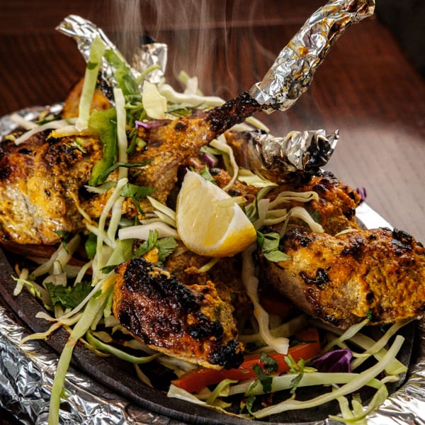 Steaming Indian chicken tikka with foil-wrapped bones, lemon, and shredded vegetables on a hot plate.