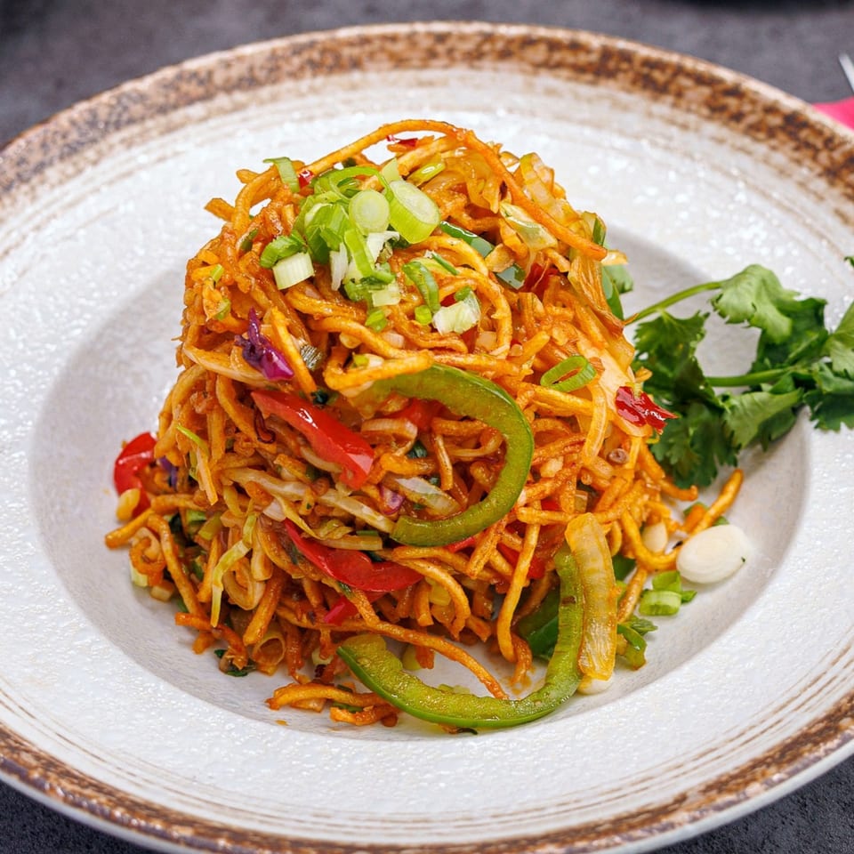 A vibrant Indian noodle dish with bell peppers, green onions, and cilantro on a white plate.