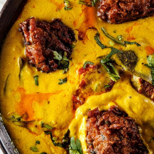 Close-up of Indian curry with dark brown pakoras in a vibrant yellow sauce, garnished with herbs.