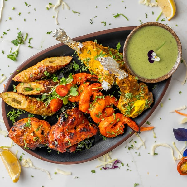 A black platter of Indian tandoori chicken, seekh kebab, and shrimp with green chutney.